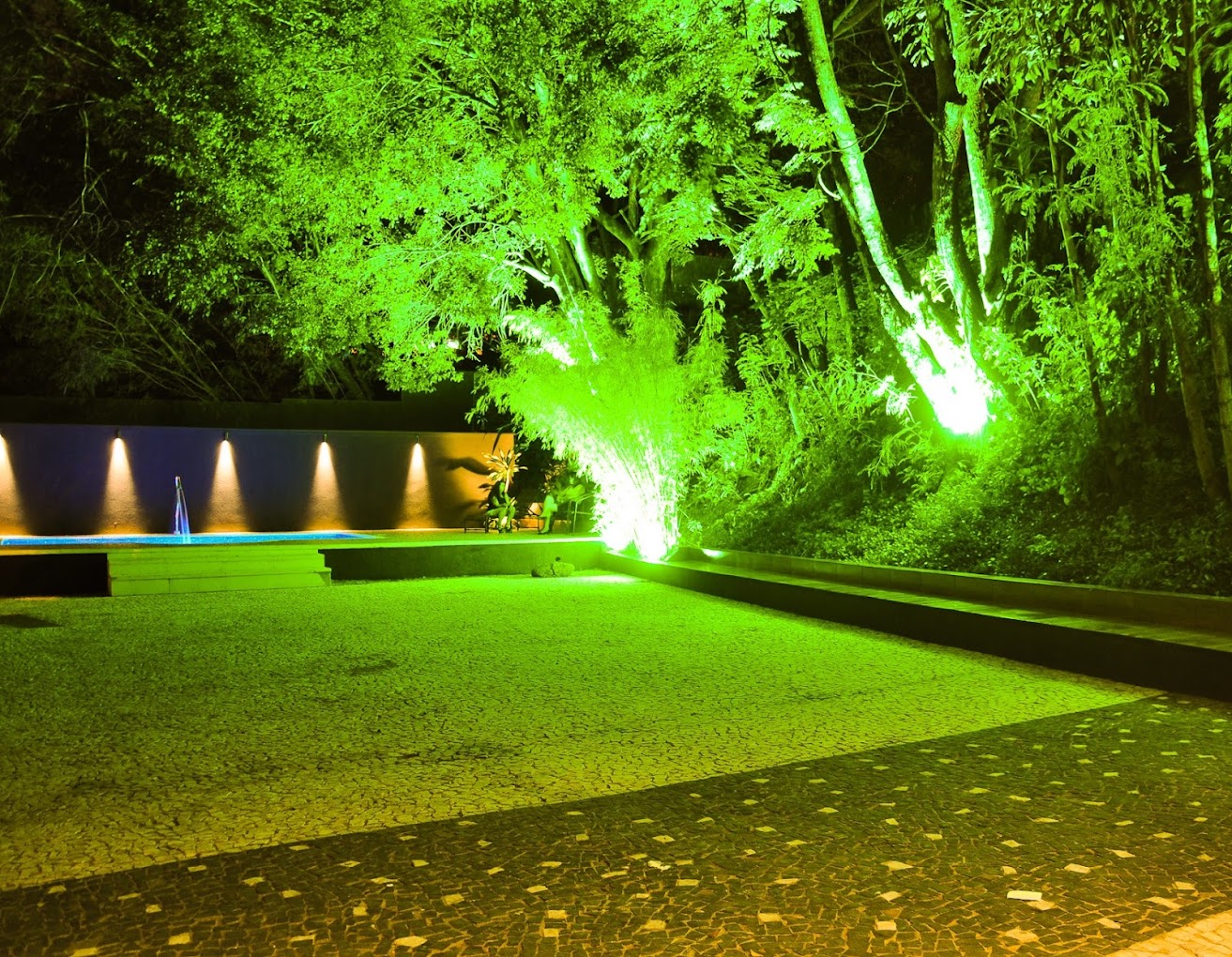 Área externa da Love Haus em Campinas, exibindo um amplo jardim gramado à noite. Grandes árvores nativas estão integradas ao paisagismo, destacadas por uma intensa iluminação cênica verde. Ao fundo, uma parede com fontes ou cascatas de água iluminadas em tons quentes de dourado e azul cria um ambiente sofisticado para festas ao ar livre.