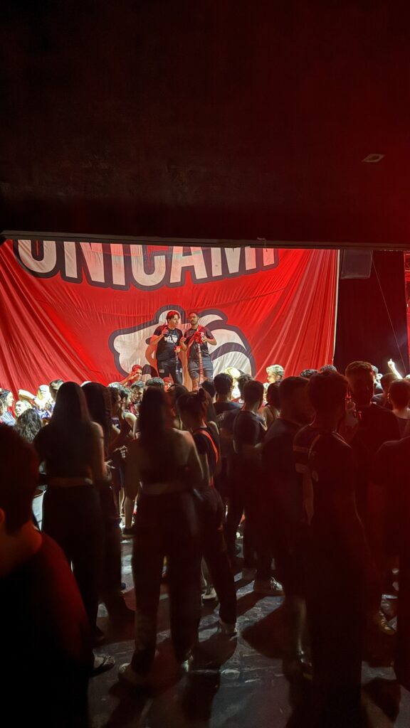 Festa universitária na Love Haus em Campinas, mostrando o palco com um grande painel vermelho com a escrita UNICAMP e o desenho de um mascote. O público jovem aparece reunido na pista de dança em frente ao palco sob iluminação vermelha e cênica intensa.