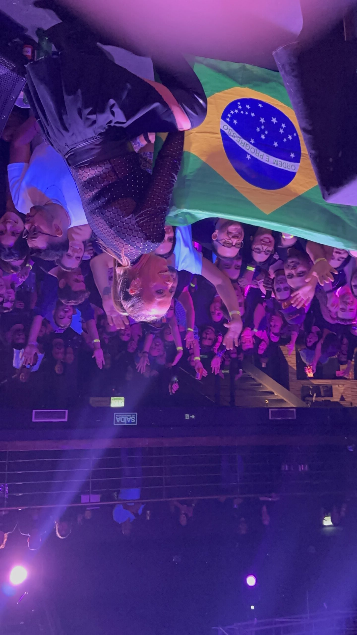 Artista se apresentando no palco da Love Haus em Campinas, agitando o público na pista de dança. A plateia aparece com as mãos para cima em clima de festa, e algumas pessoas na frente seguram a bandeira do Brasil. O ambiente está lotado e iluminado com luzes cênicas em tons de roxo e rosa, com o público do mezanino visível ao fundo.