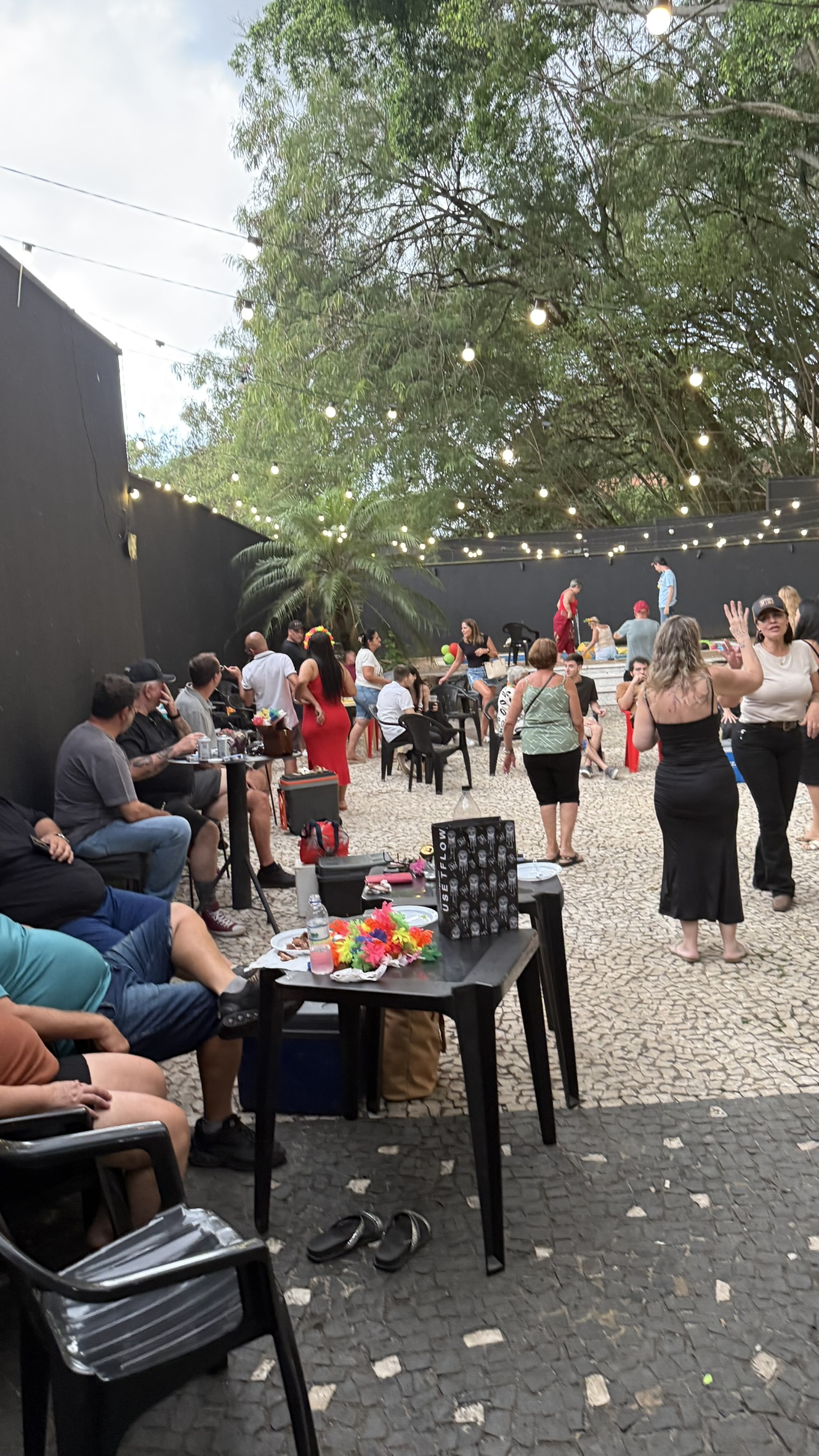 Público reunido na área externa da Love Haus em Campinas durante um evento diurno de tarde. À esquerda, pessoas aparecem sentadas em mesas e lounges de apoio conversando, enquanto ao fundo outros convidados circulam pelo espaço plano cercado por muros escuros e árvores. Acima, um varal de luzes decorativas (string lights) está estendido entre as árvores, criando uma ambientação de festa ao ar livre.