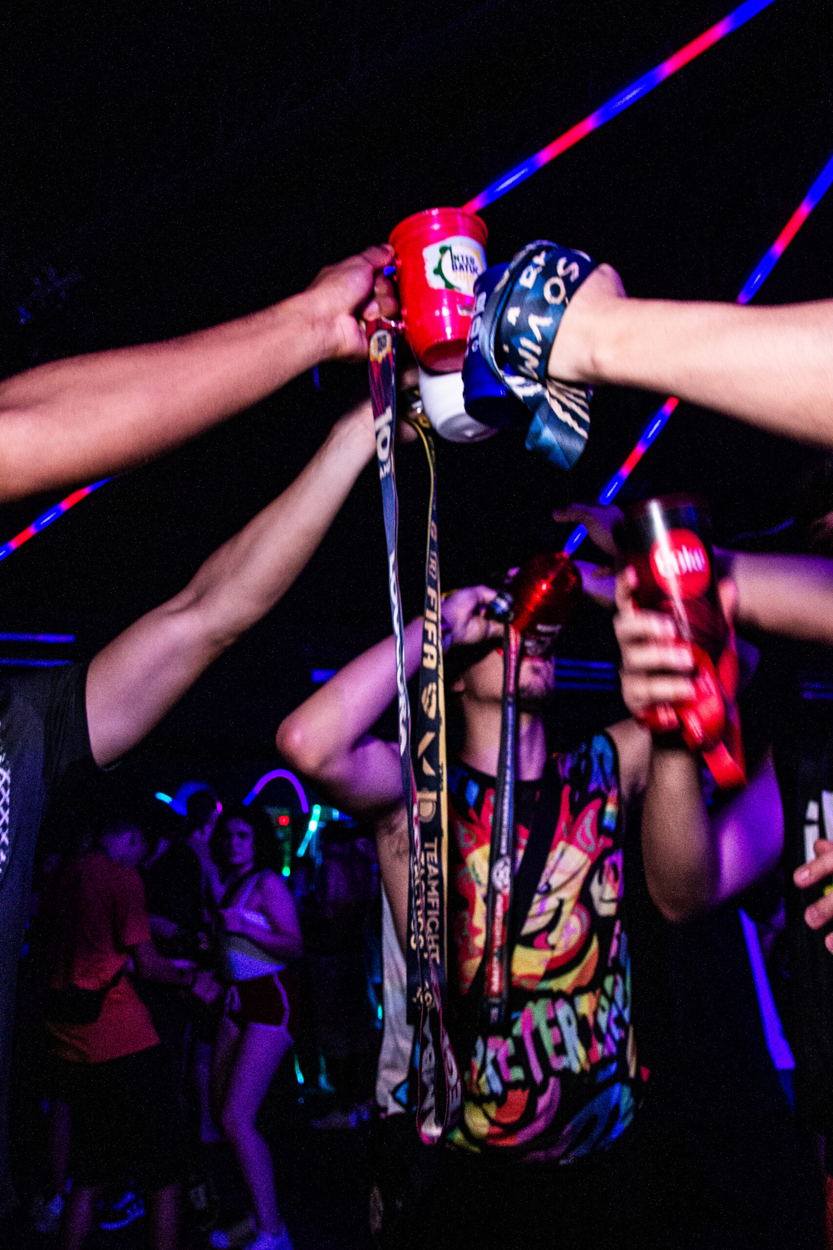 Jovens erguendo copos e canecas coloridas em um brinde coletivo na pista de dança da Love Haus em Campinas. É possível ver canecas com tirantes de atléticas universitárias levantadas em direção ao teto, que possui linhas de iluminação neon em tons de azul e rosa. No centro, um jovem com regata colorida estampada curte a balada em um ambiente animado e com luz cênica escura.