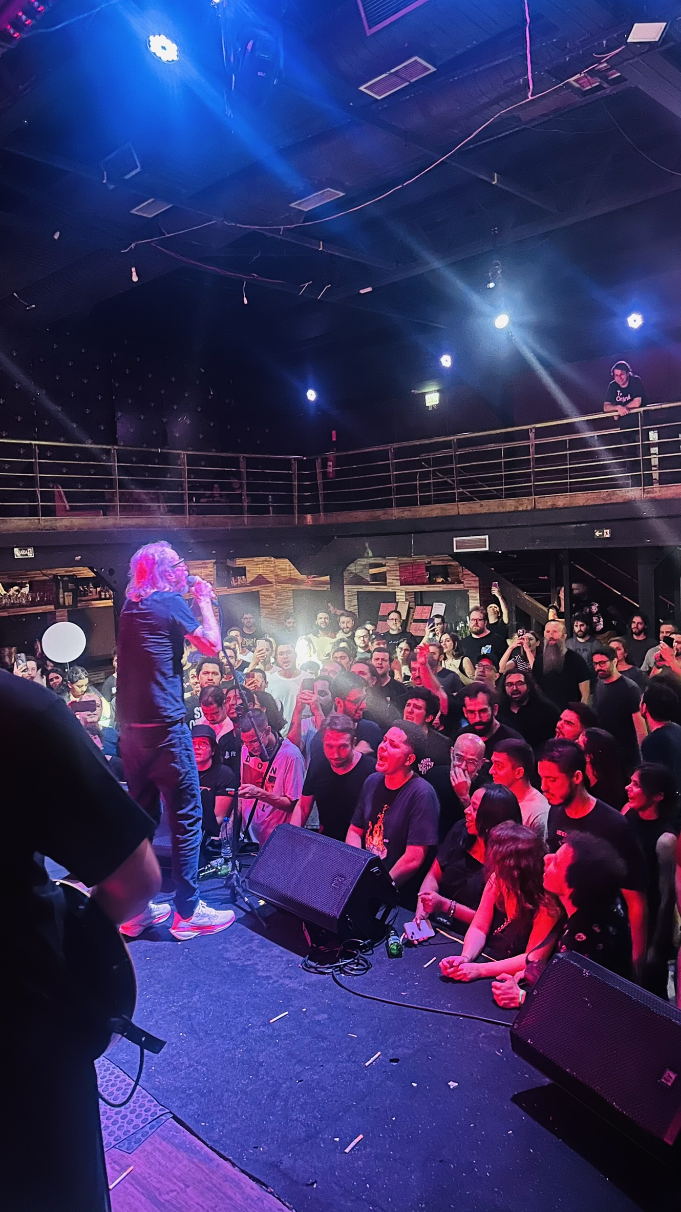 Cantor ou MC se apresentando de perfil no palco iluminado da Love Haus em Campinas, segurando o microfone sob forte luz branca cênica. À sua frente, o público lota a pista de dança com iluminação em tons de rosa e vermelho. Ao fundo, destaca-se a estrutura metálica do mezanino superior de dois andares com guarda-corpo e caixas de som profissionais suspensas.