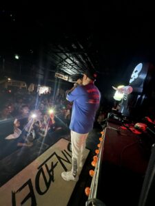 Visão lateral do palco da Love Haus em Campinas durante a apresentação de um artista de trap ou funk, vestindo jaqueta azul e calça branca, cantando com microfone na mão. À sua frente, o público lota a pista de dança com as lanternas dos celulares acesas, criando um efeito de luzes brilhantes. O palco conta com spots de iluminação amarela na borda.