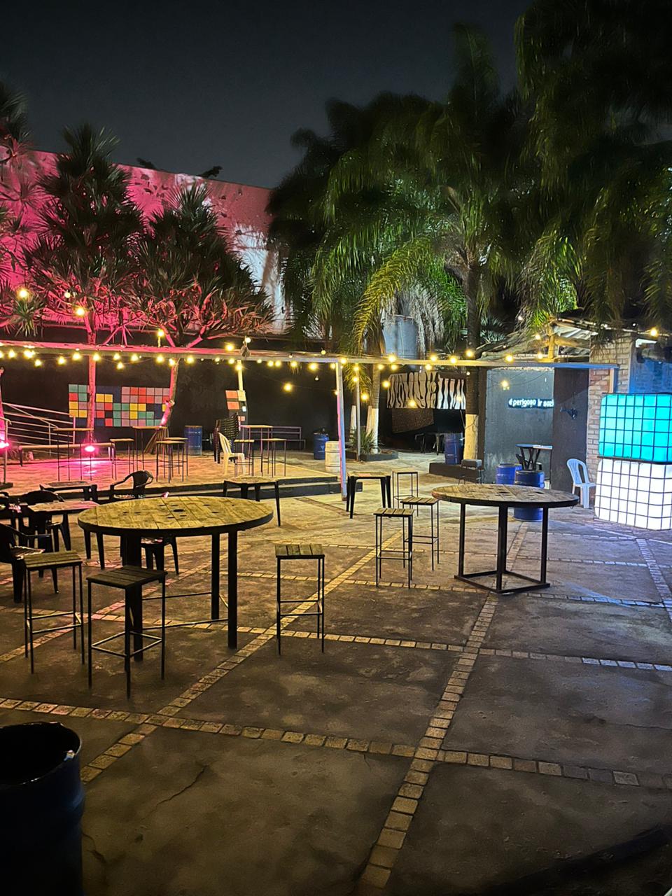 Área externa de convivência da Chácara Love Haus à noite, exibindo um pátio pavimentado com mesas bistrô redondas de madeira e banquetas altas. O ambiente é decorado com varais de lâmpadas suspensas de luz quente, com palmeiras e árvores ao fundo recebendo iluminação cênica verde e rosa, além de um telão de LED posicionado na lateral direita.