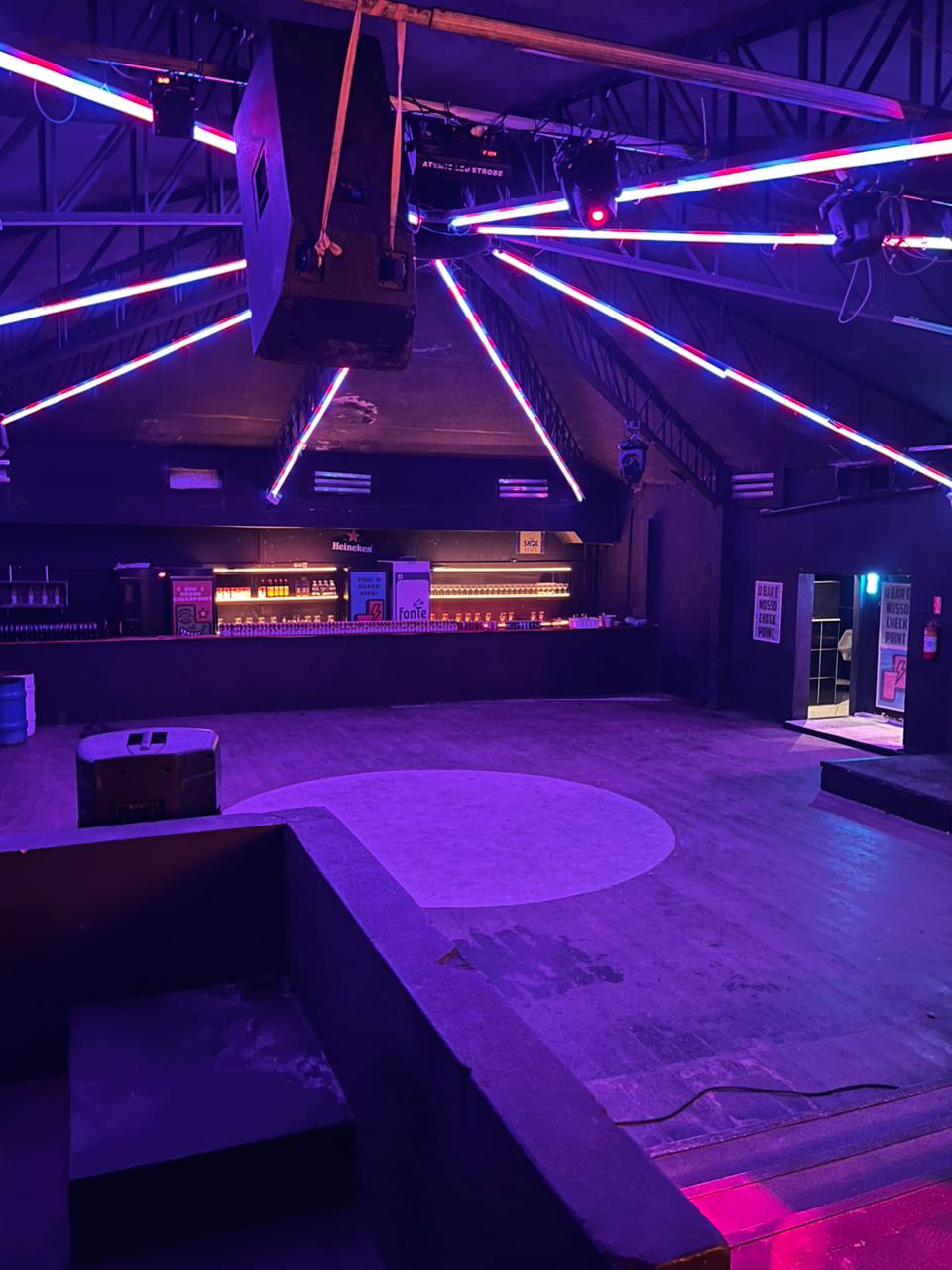 Vista interna da pista de dança do Espaço Tetriz e Love Haus em Campinas, destacando o teto arquitetônico inclinado em formato de pirâmide. Longas barras de LED neon brilham em tons de azul, rosa e vermelho, estendendo-se do centro do teto até as laterais da pista como raios. Ao fundo, o balcão de bar aparece iluminado em tom quente, com caixas de som profissionais suspensas e lounges em primeiro plano sob luz ultravioleta.