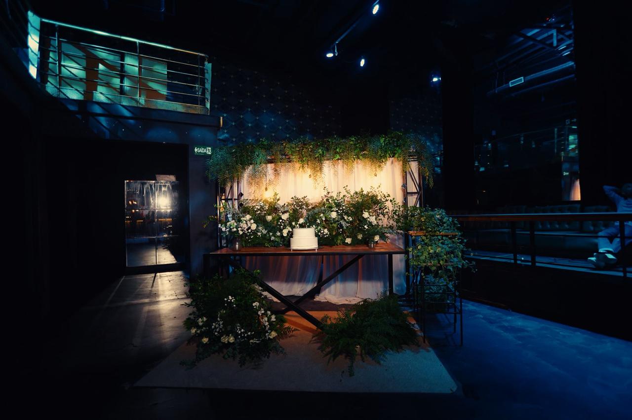 Lounge decorado e instagramável no interior da Love Haus em Campinas. O espaço conta com uma mesa rústica central de madeira, arranjos de plantas tropicais verdes e samambaias suspensas, iluminadas por uma parede de luzes quentes amareladas ao fundo. O ambiente ao redor possui iluminação neon azul cênica e telas digitais.