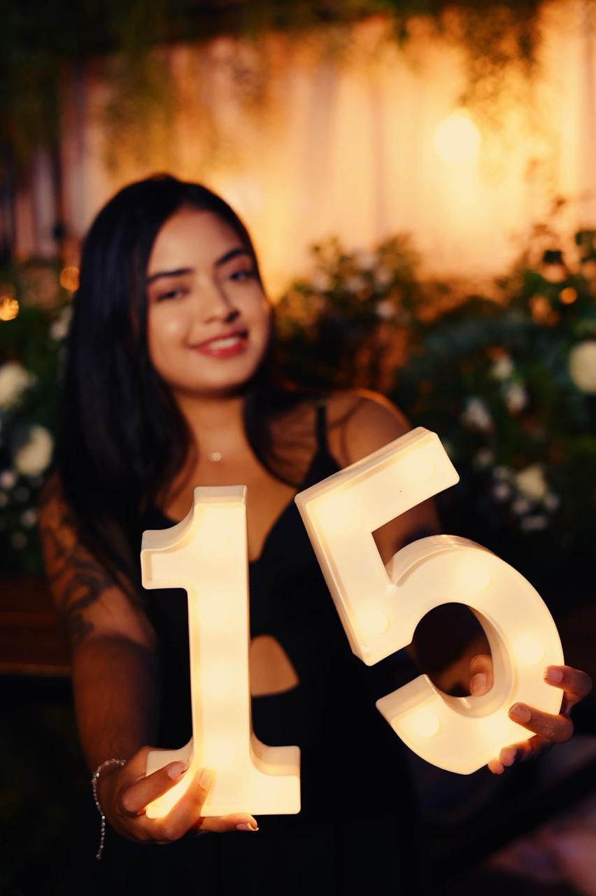 Jovem sorridente segurando letreiros luminosos em formato do número 15 no espaço decorado da Love Haus em Campinas. Ao fundo, uma mesa rústica de madeira com arranjos de flores brancas, folhagens verdes e cortina de luzes quentes (luzes de LED de filamento), criando um cenário elegante para festa de debutante ou aniversário.