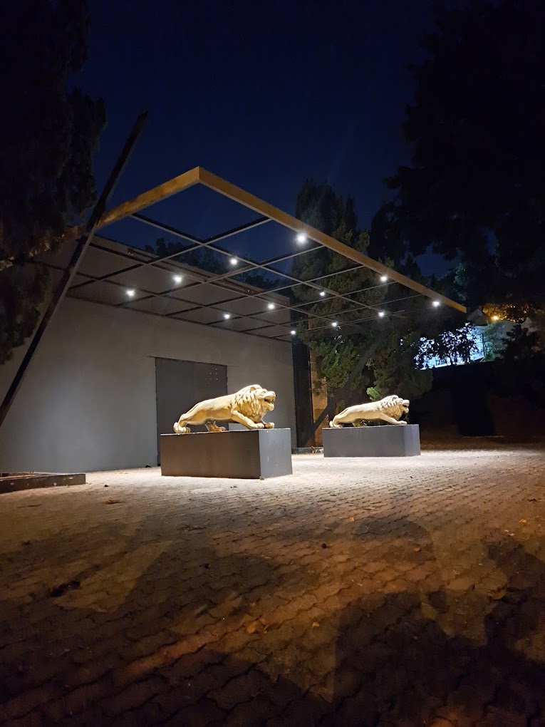 Fachada da Love Haus em Campinas durante a noite, destacando a entrada principal com piso de paralelepípedo iluminado. Há duas grandes estátuas de leões dourados imponentes sobre pedestais escuros na recepção. Acima, uma estrutura de pergolado metálico moderno com varal de luzes quentes estilo gambiarra (luzes de quermesse) ilumina o acesso sob o céu noturno.